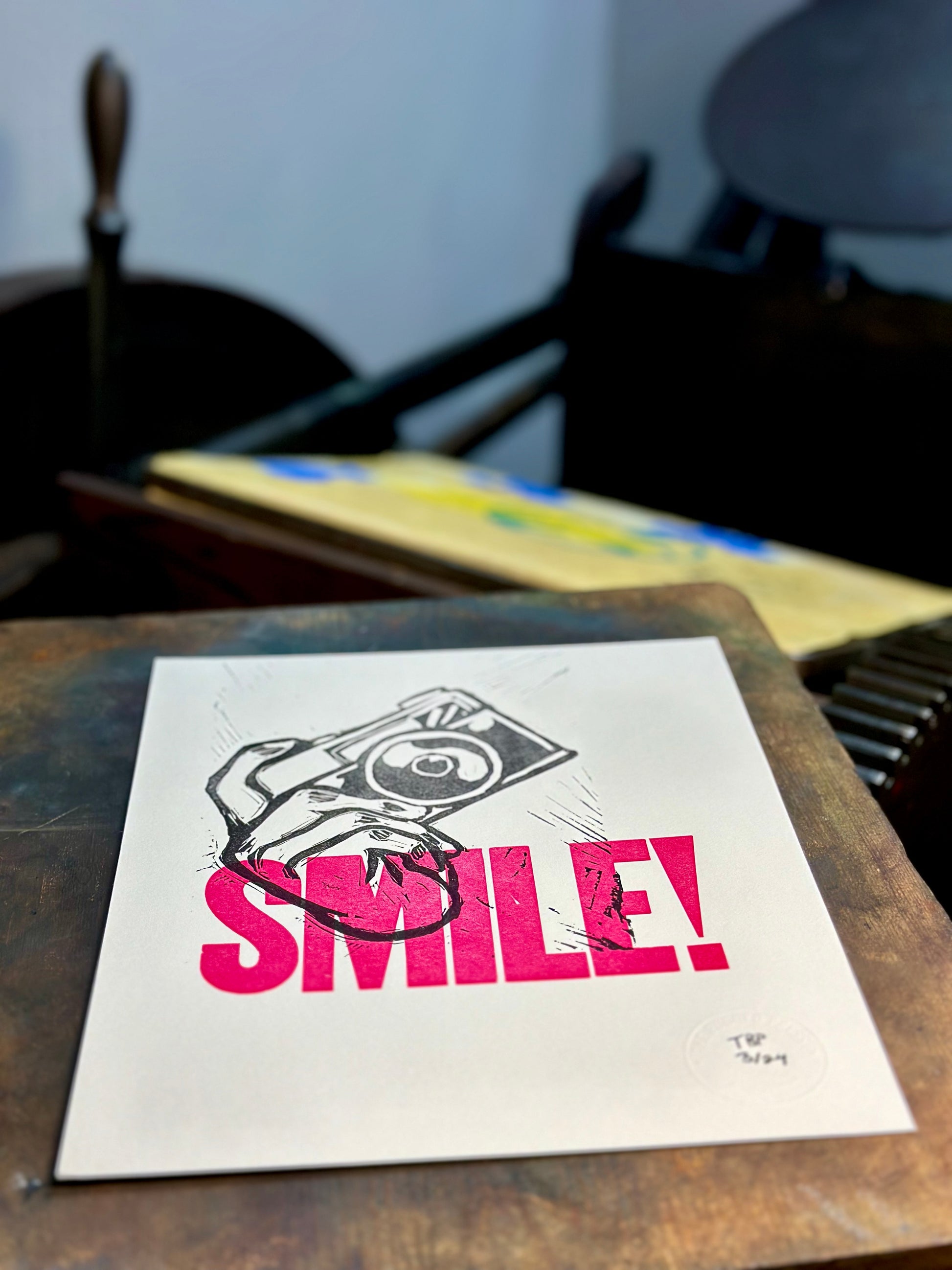 Aperture Priority handmade letterpress art print. Features the word "smile" in pink ink with a carved hand holding a camera. Press view.