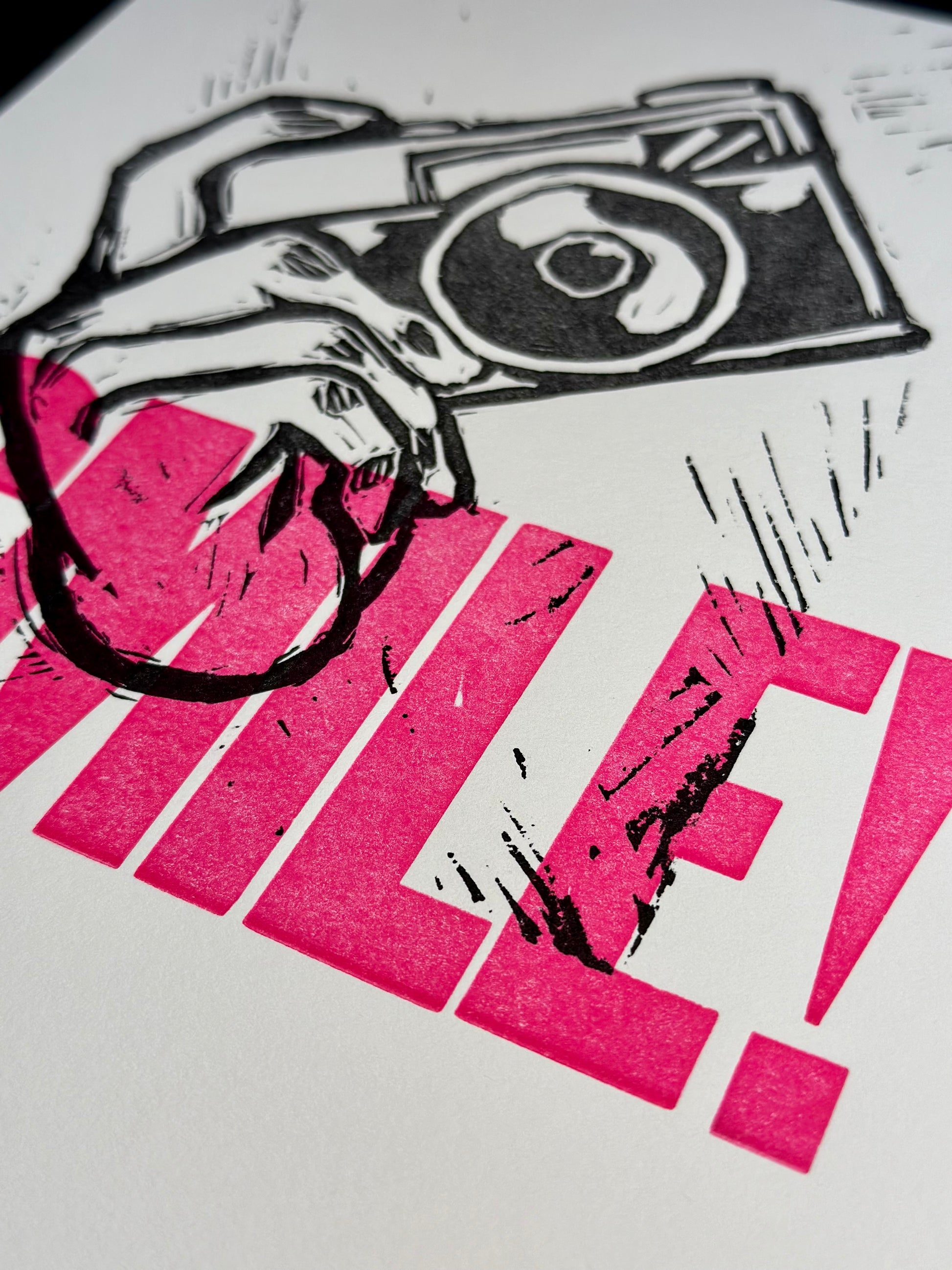 Aperture Priority handmade letterpress art print. Features the word "smile" in pink ink with a carved hand holding a camera. Detail view.