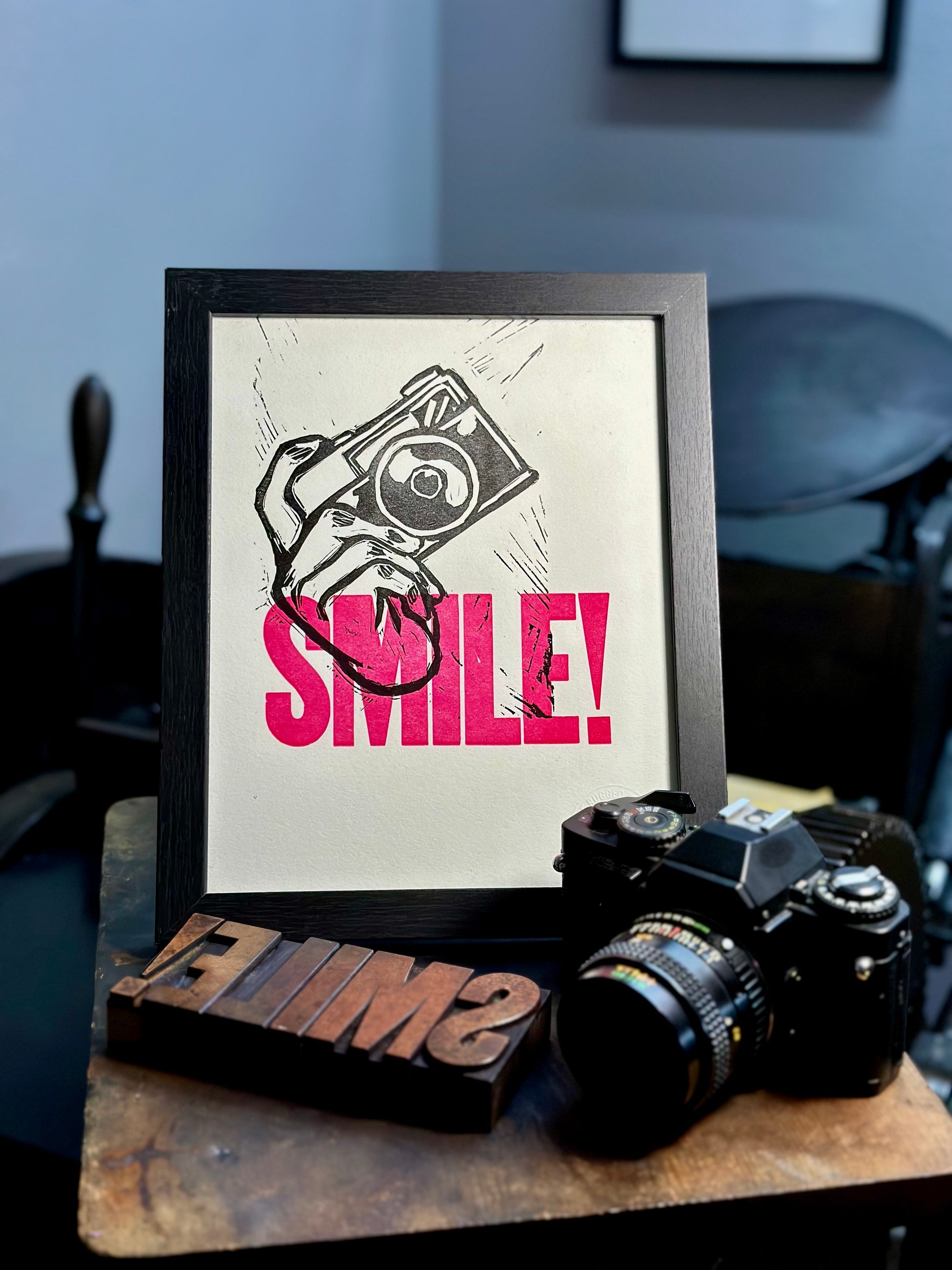 Aperture Priority handmade letterpress art print. Features the word "smile" in pink ink with a carved hand holding a camera. Framed view with printing press in background.