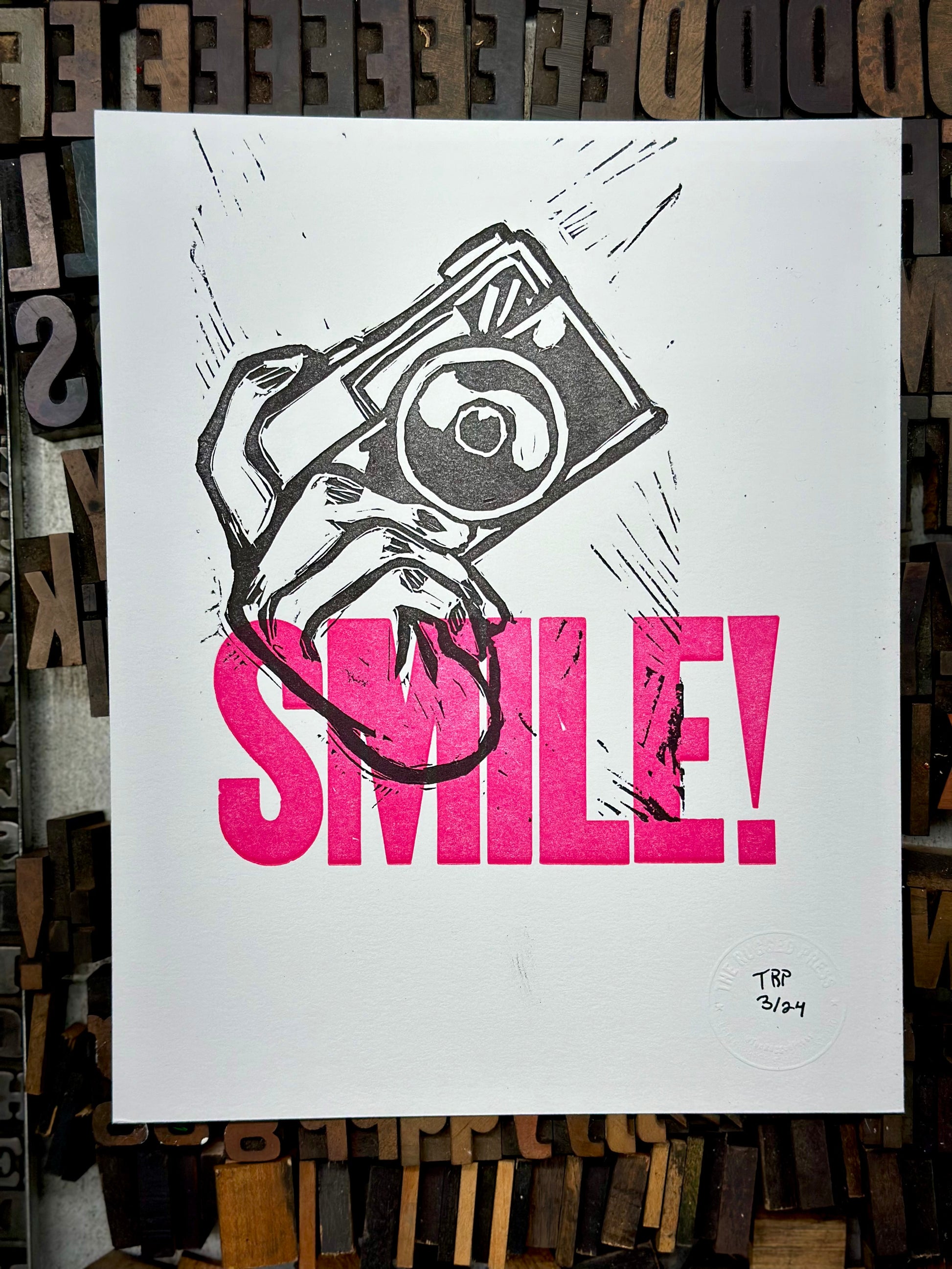 Aperture Priority handmade letterpress art print. Features the word "smile" in pink ink with a carved hand holding a camera. Print placed on historic wood type view.