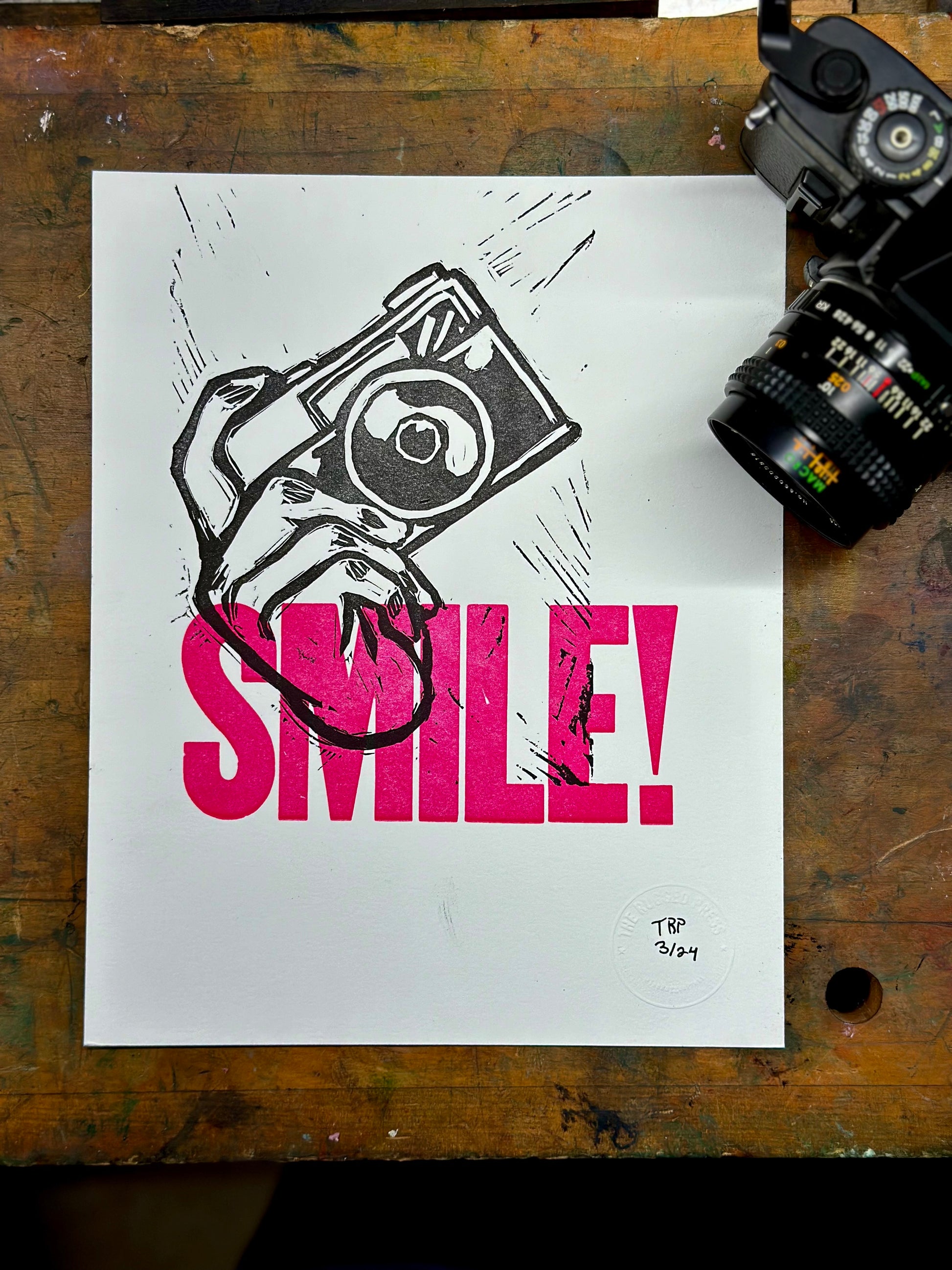 Aperture Priority handmade letterpress art print. Features the word "smile" in pink ink with a carved hand holding a camera. unframed view with camera.